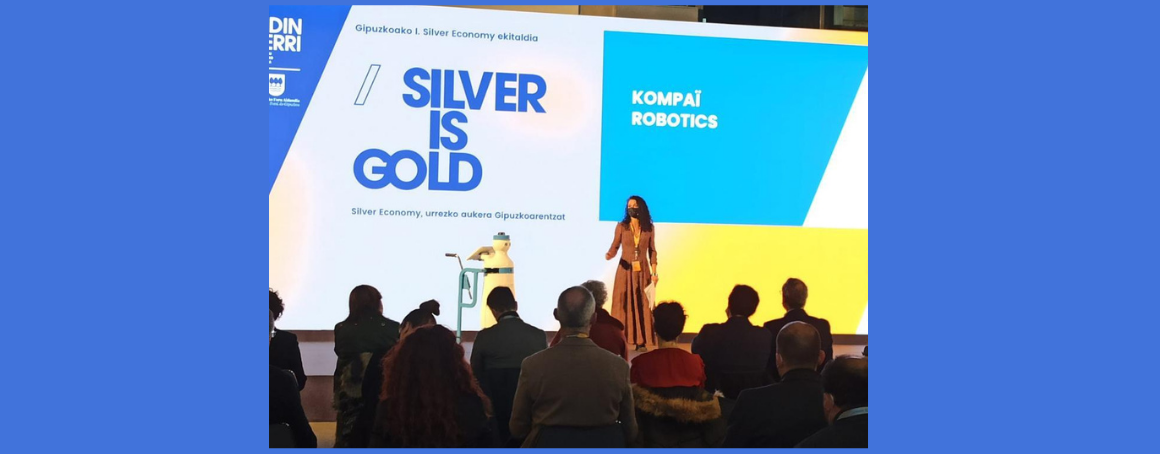 Image of a woman on stage with the text "silver is gold" and Kompai Robotics on the screen behind.