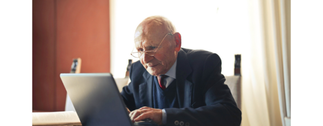 Older white male, wearing glasses and a navy suit, using a laptop.