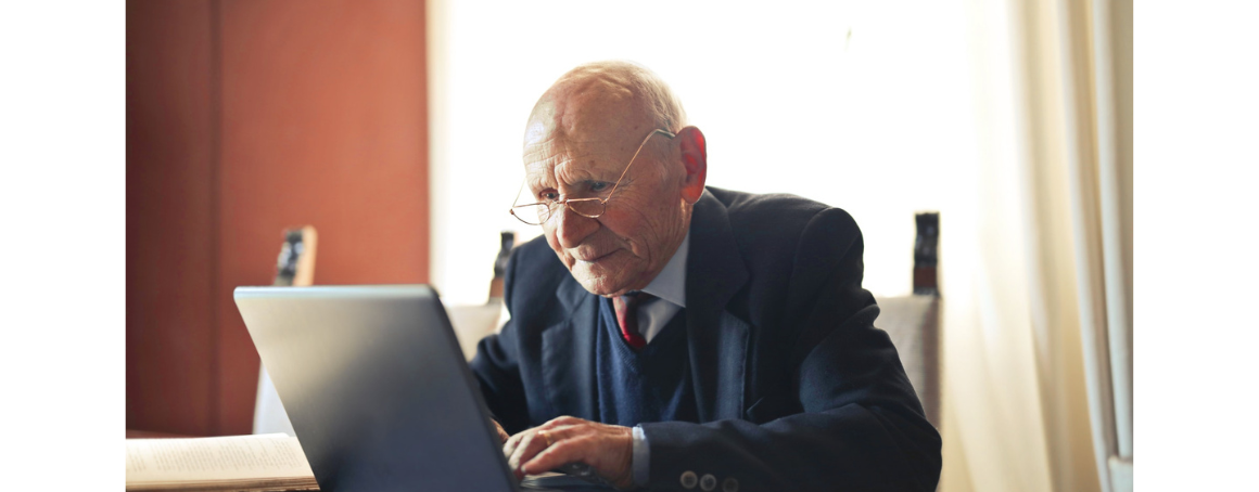 Older white male, wearing glasses and a navy suit, using a laptop.