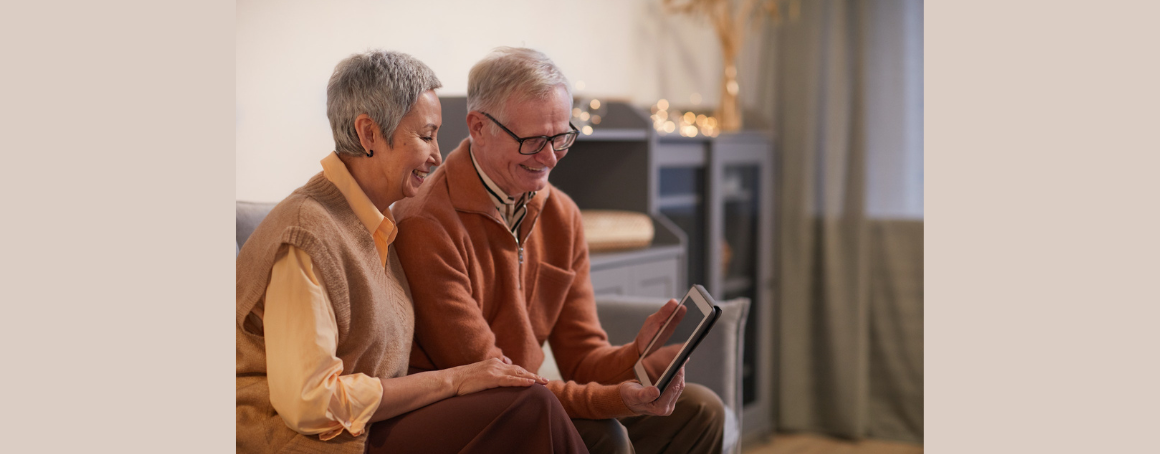 Two older white adults sitting on a couch, smiling using a digital tablet.