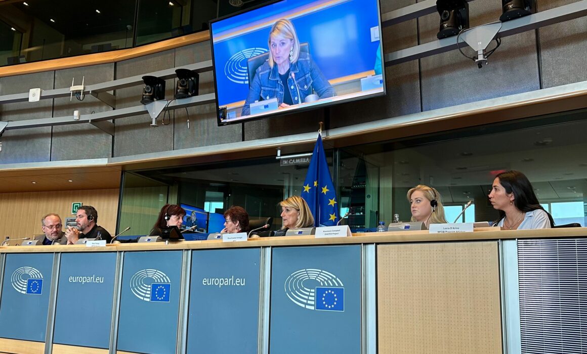 View of speakers at the European Parliament event. It shows two men on the left side and 5 women sitting next to each other behind a table. Behind them, a screen showing a blonde woman.
