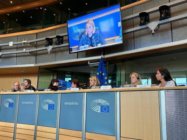 View of speakers at the European Parliament event. It shows two men on the left side and 5 women sitting next to each other behind a table. Behind them, a screen showing a blonde woman.