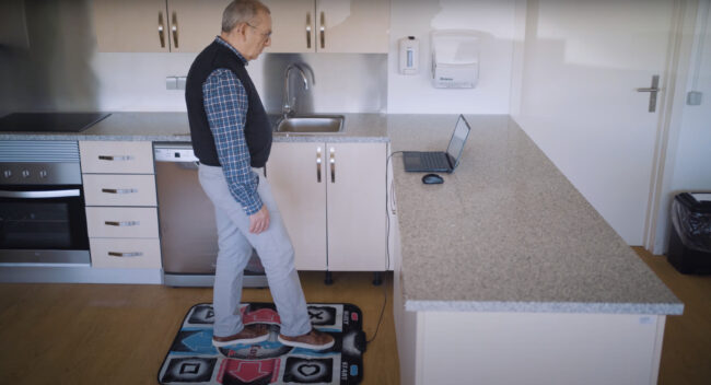 In the image you can see an elderly man in a kitchen, in front of a computer and on the DanceMove mat, a digital solution developed by the SHAPES project.